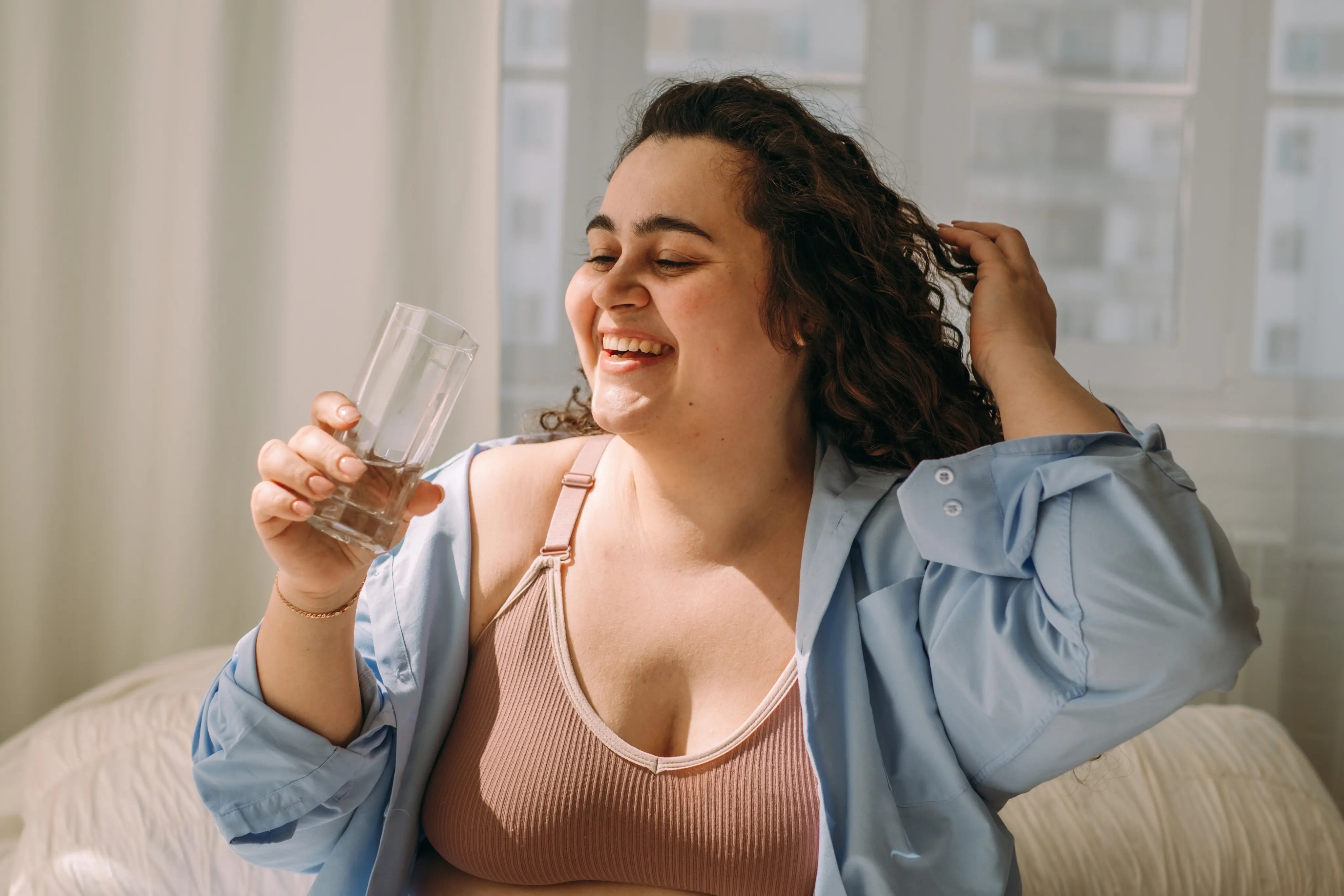 Smiling woman with curly hair in a bra and open shirt holding a glass of water indoors.