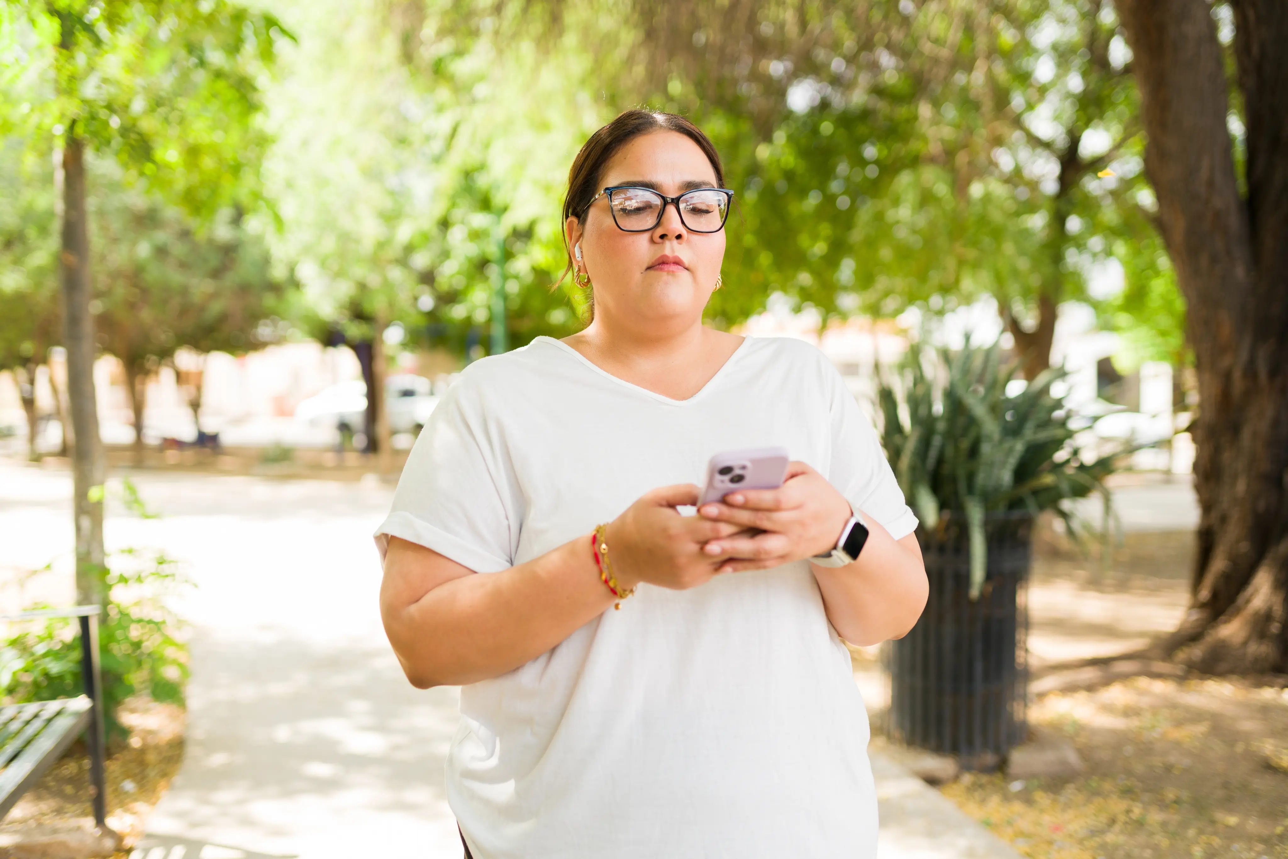 Woman wearing glasses and a white shirt using a smartphone outdoors with trees in the background.