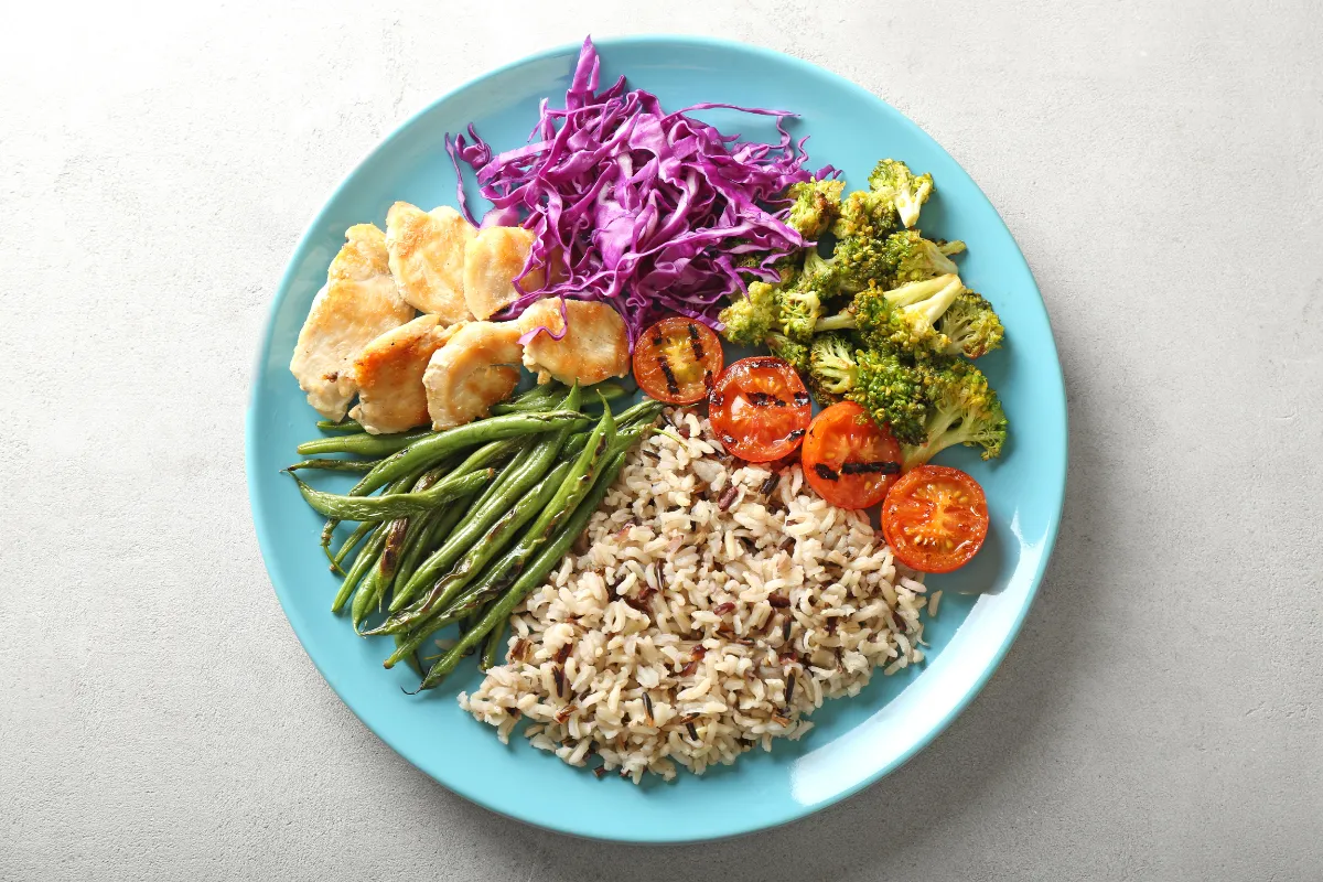 Blue plate with grilled chicken, green beans, shredded purple cabbage, roasted broccoli, grilled tomatoes, and mixed rice.