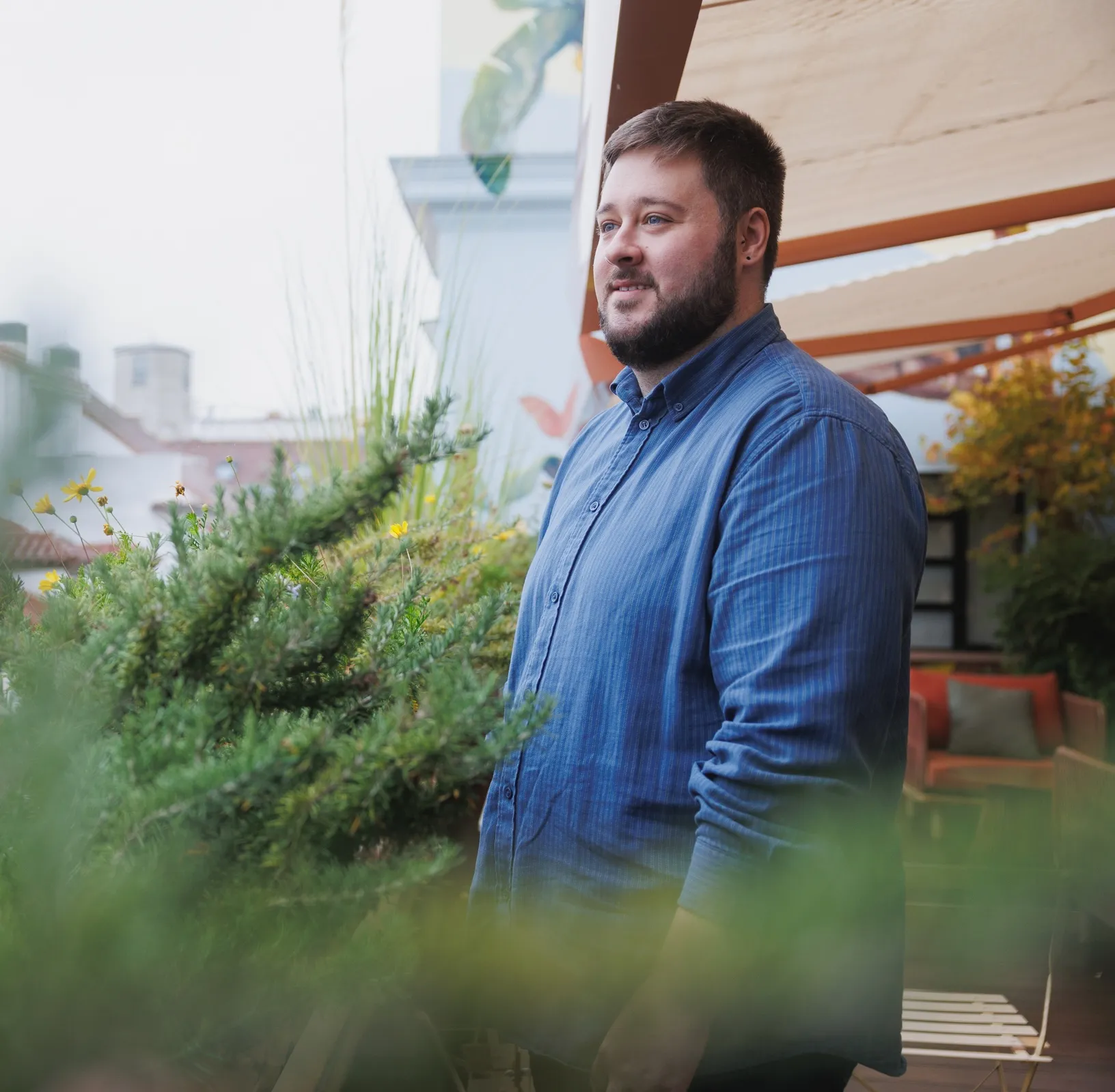 Smiling bearded man in a blue shirt standing near plants on a patio with outdoor seating.