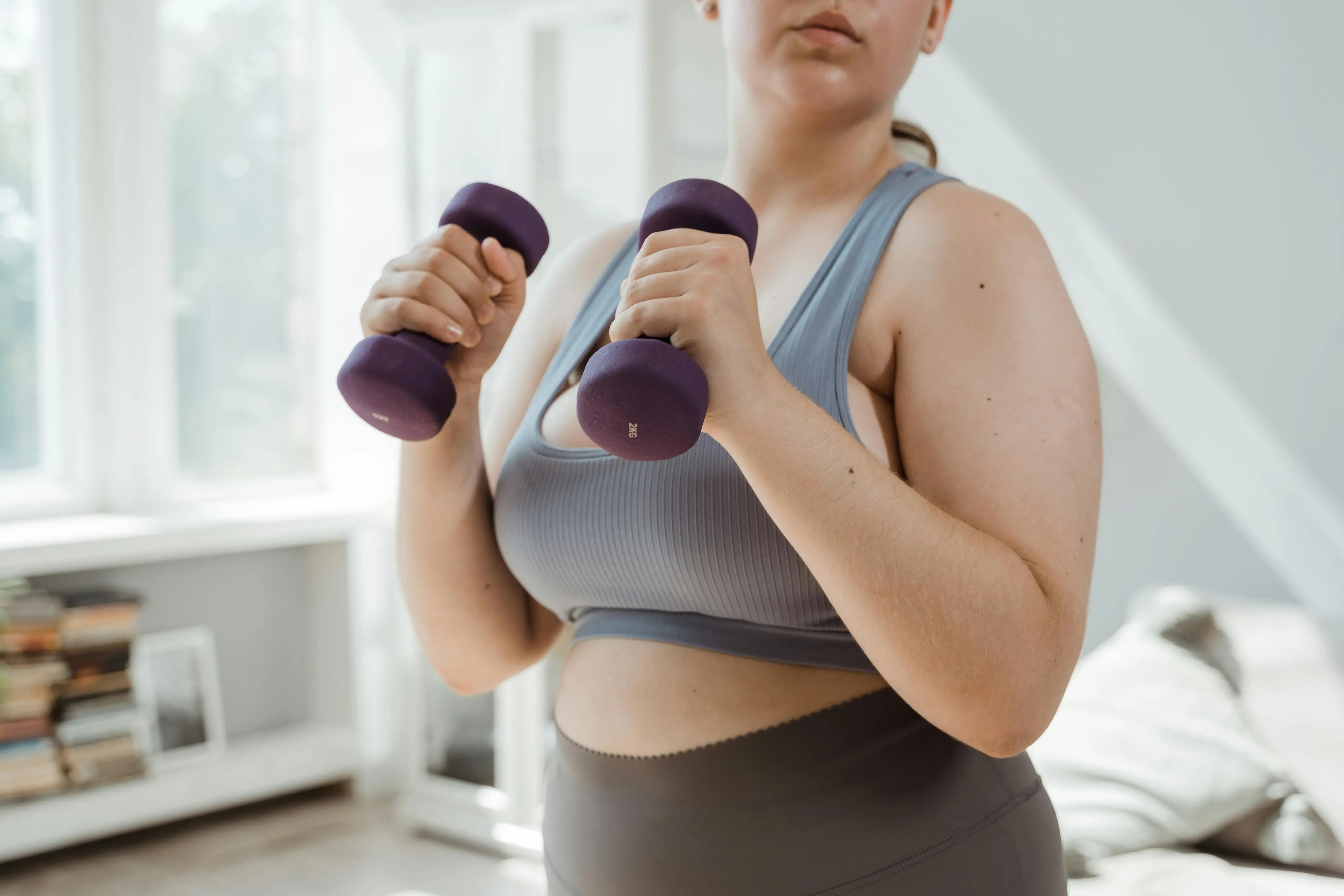 Woman in gray workout clothes lifting purple dumbbells indoors near window.