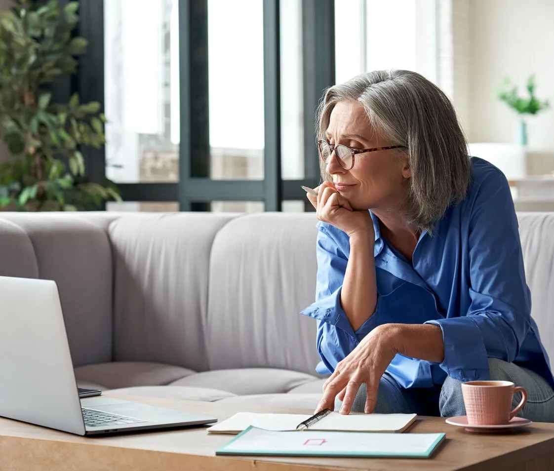 Mature woman in glasses sitting on couch, working with laptop and notebook on coffee table.