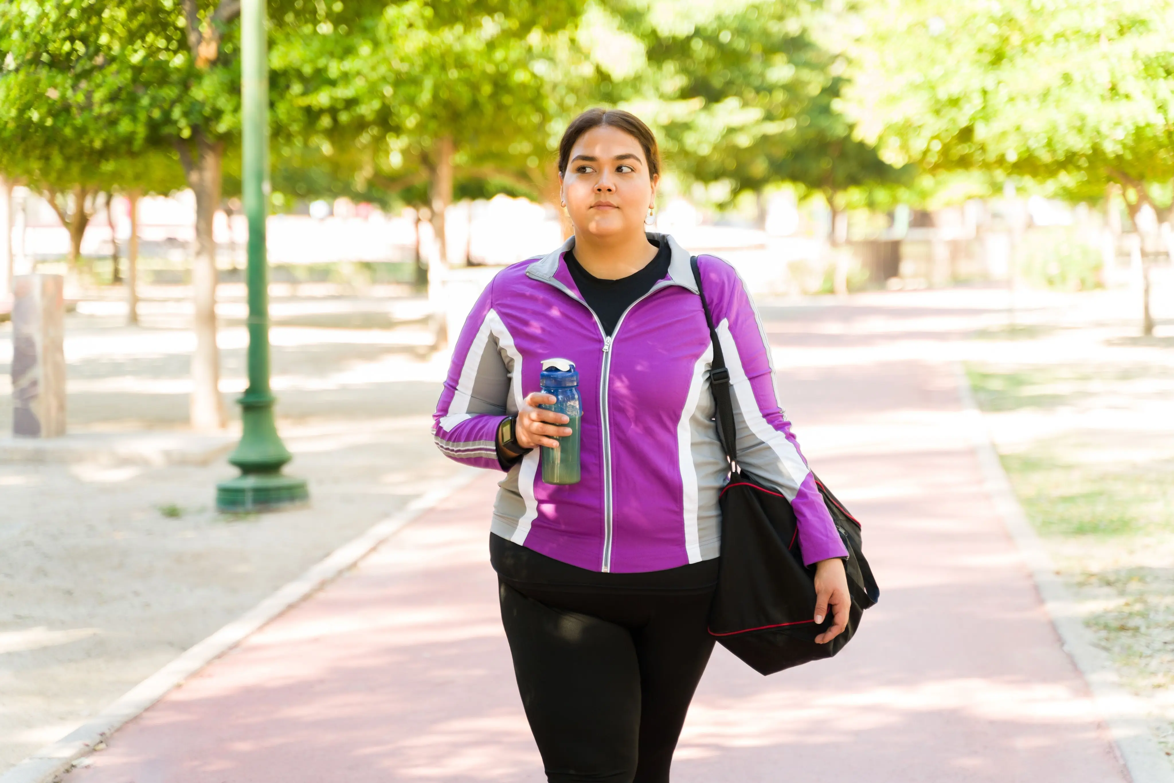 Woman in a purple jacket walking on a tree-lined path holding a water bottle and a black gym bag.