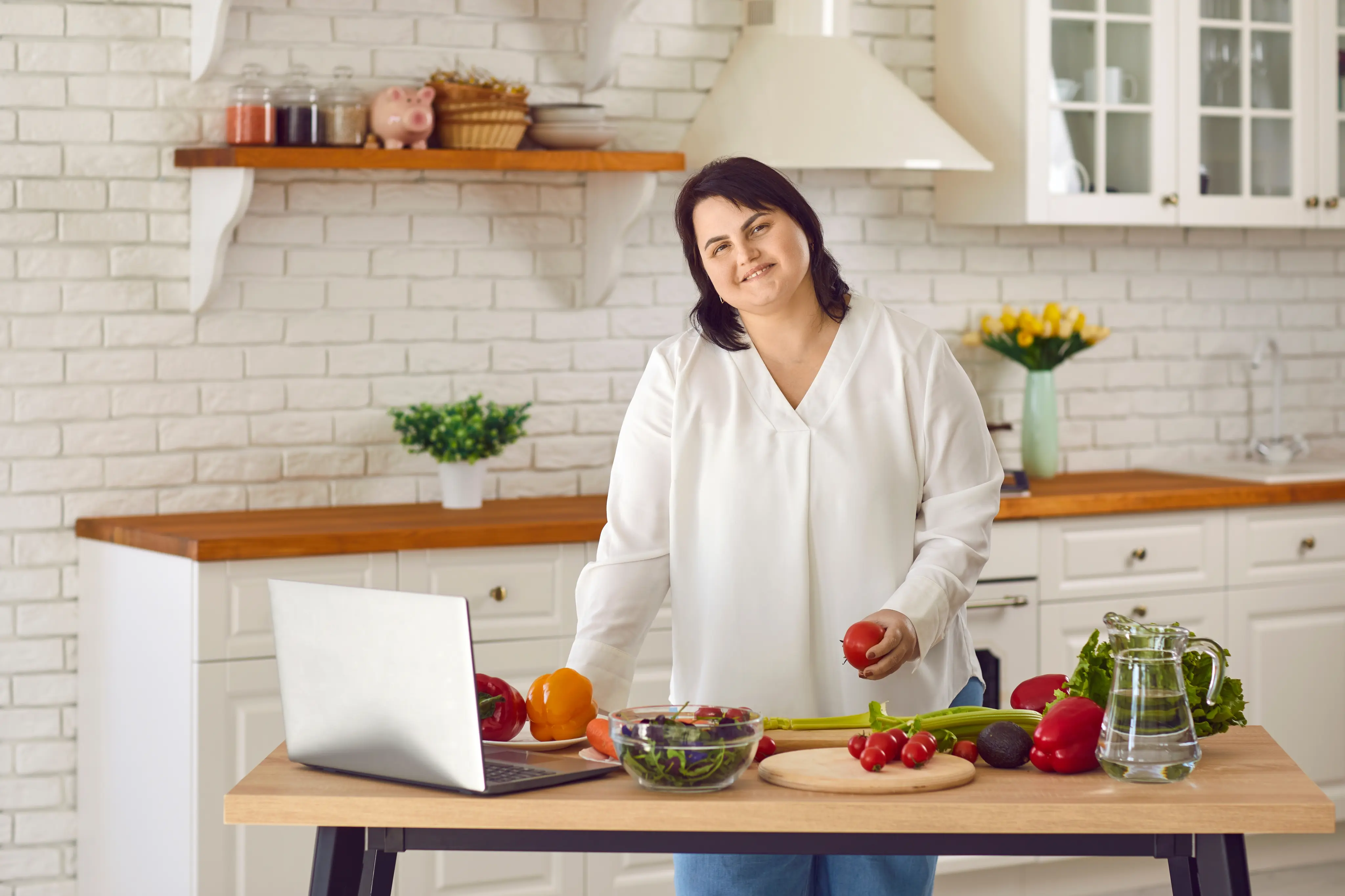 Woman in white blouse cooking with vegetables and laptop on a kitchen table.