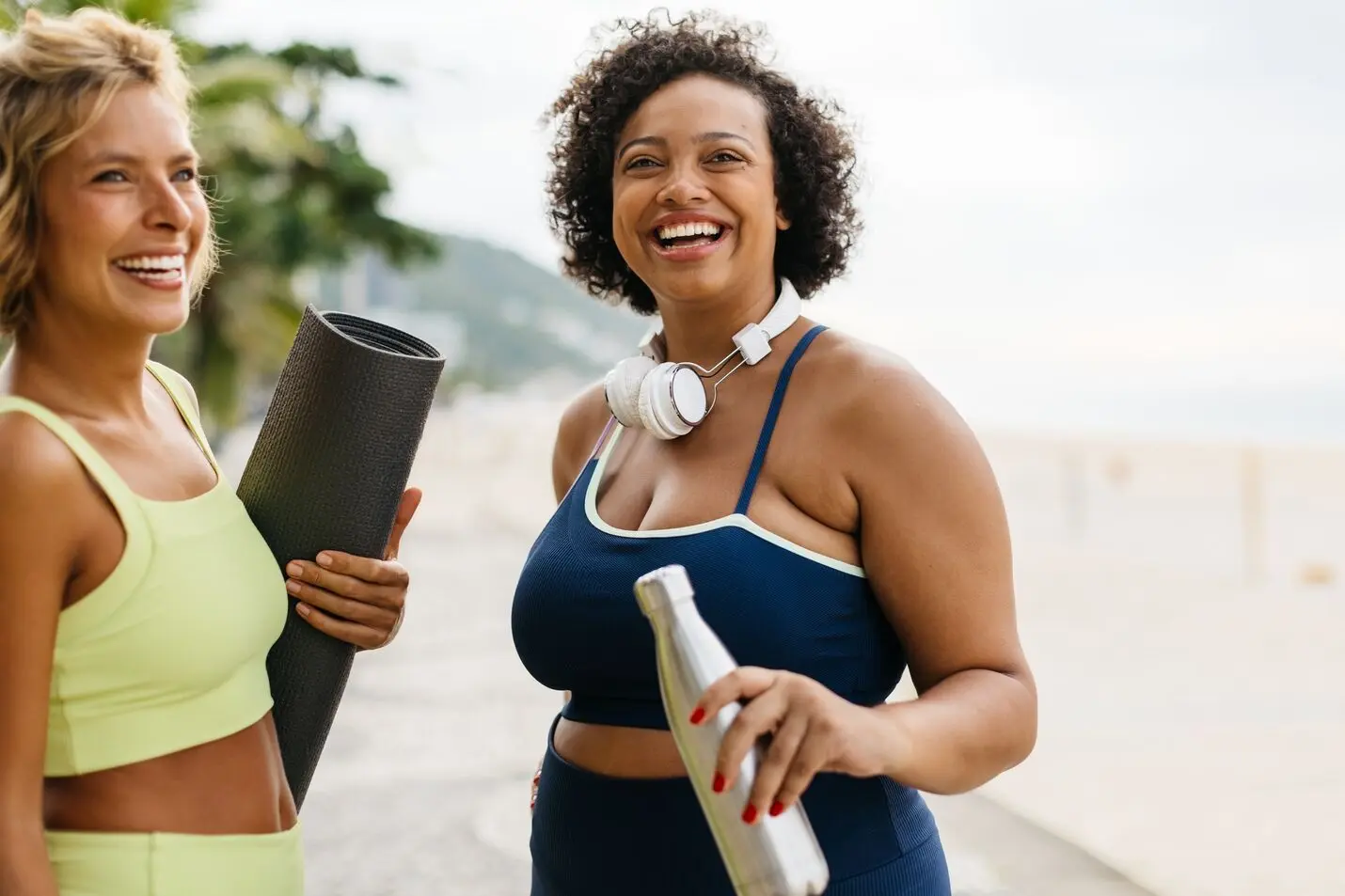 Two women in workout clothes smiling outdoors; one holds a yoga mat, the other a water bottle and headphones.