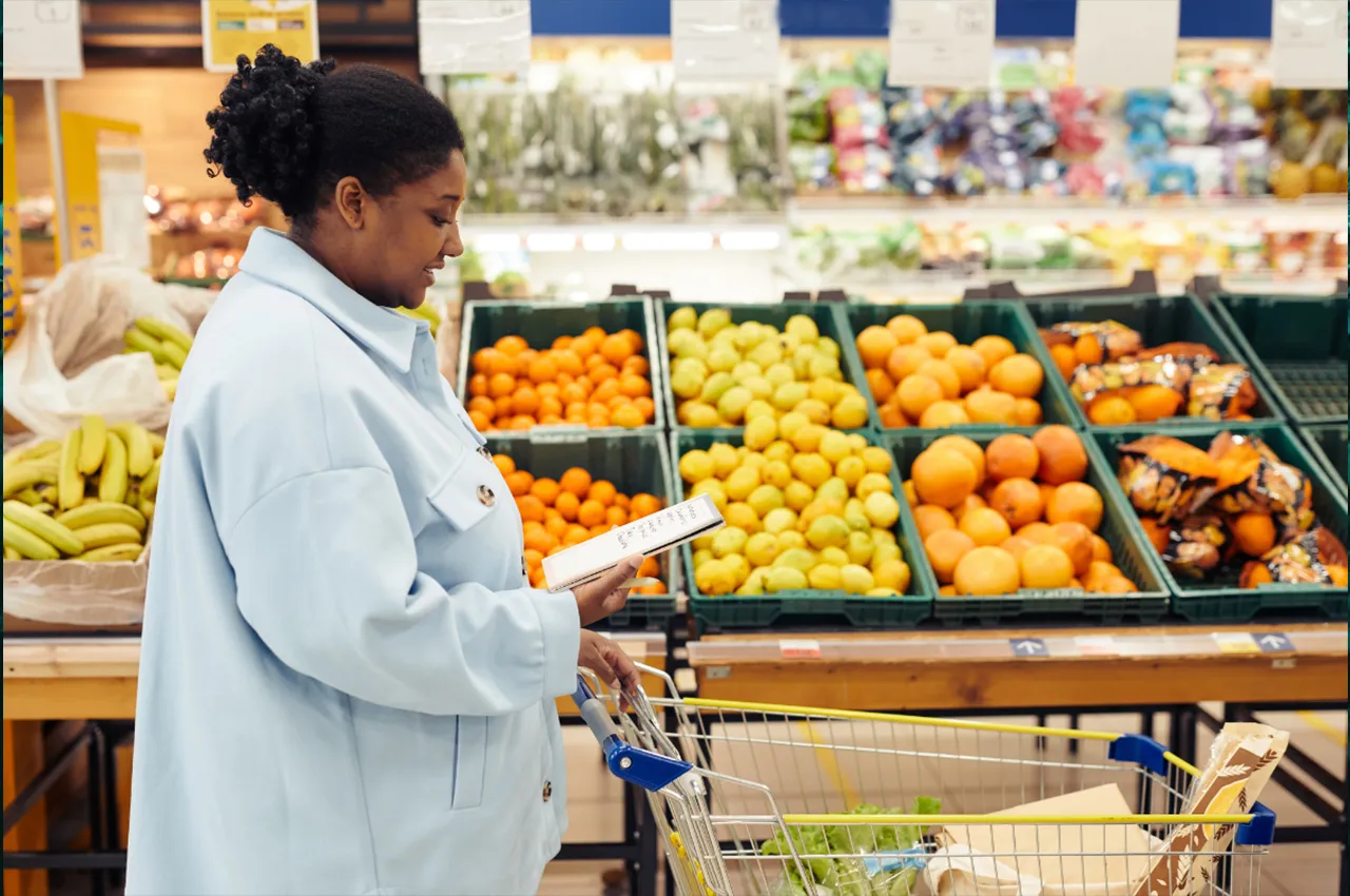 Woman in a light blue jacket checking a grocery list while shopping in a supermarket produce aisle.