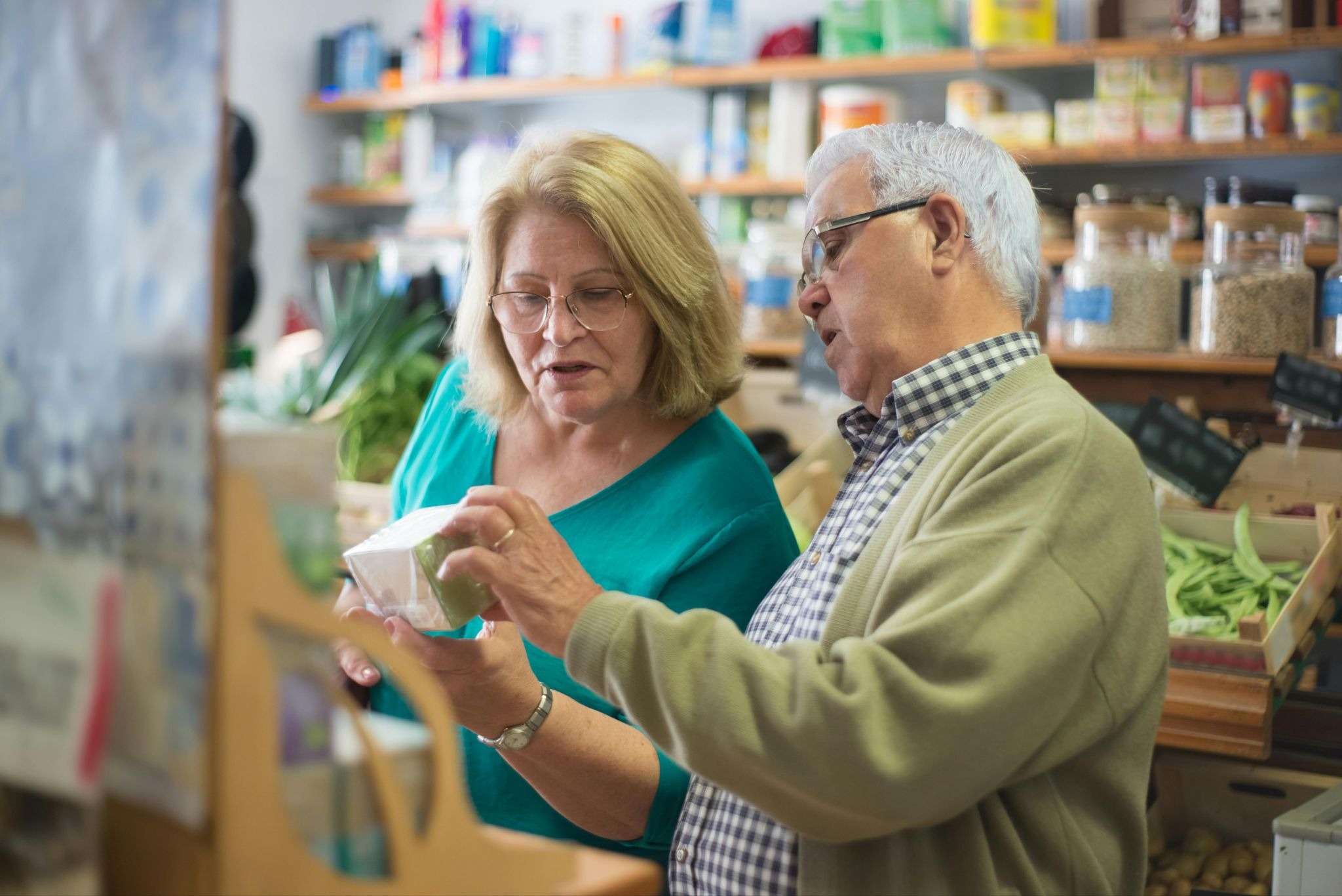A senior man and woman wearing glasses examine a product label together in a small grocery store or pharmacy.