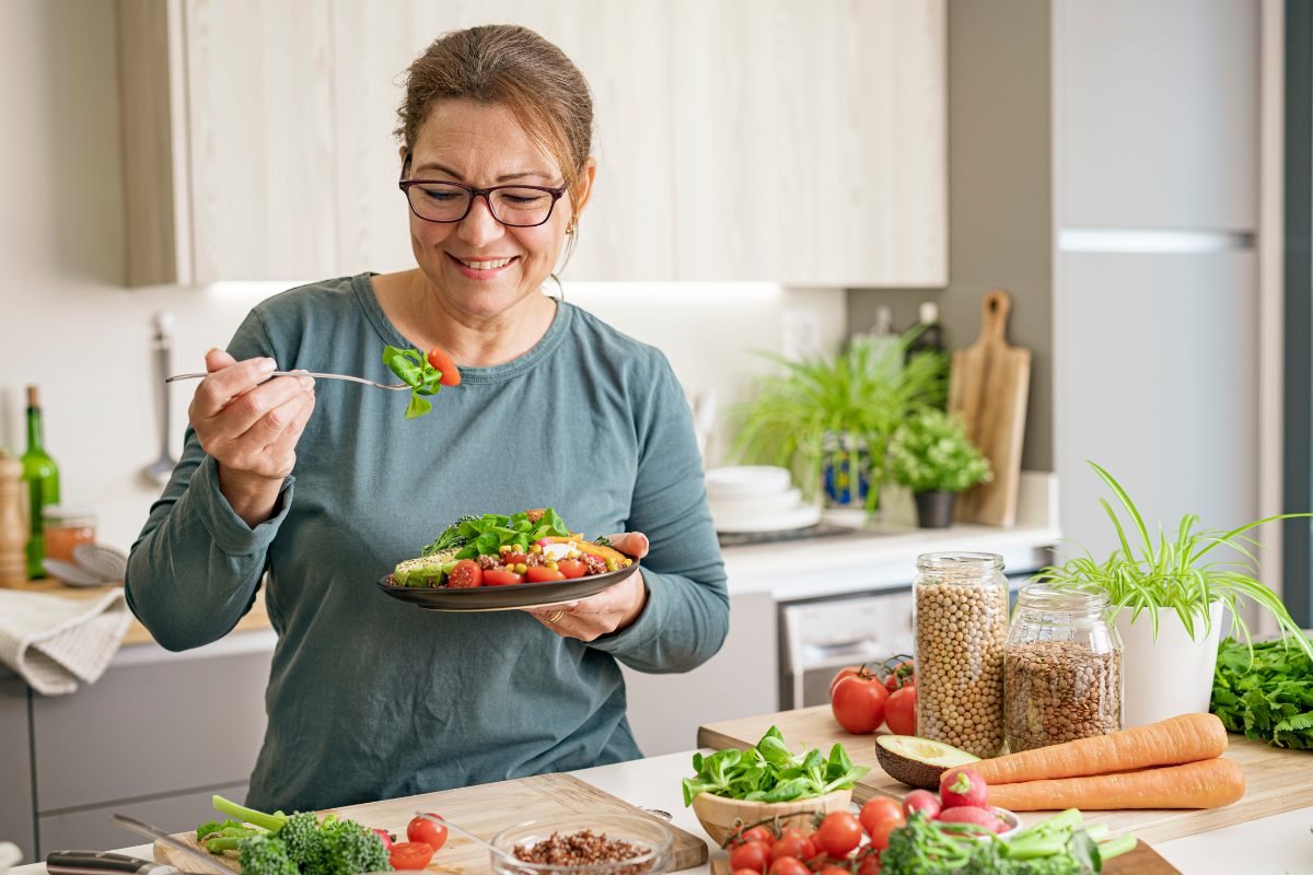 A woman enjoying a healthy meal in a bright kitchen surrounded by fresh vegetables, lentils, and chickpeas.