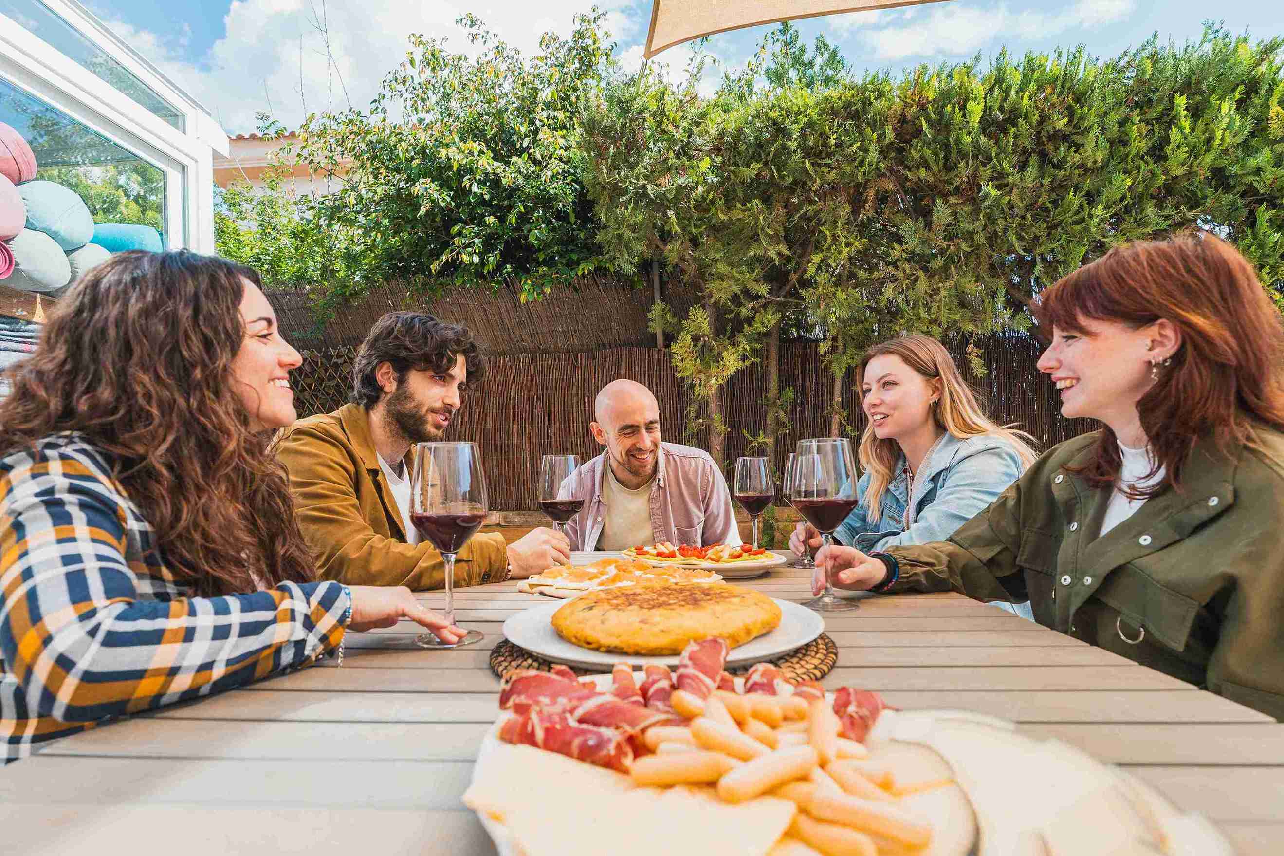 Un grupo de amigos está sentado alrededor de una mesa al aire libre, compartiendo comida y vino en un ambiente agradable y relajado.