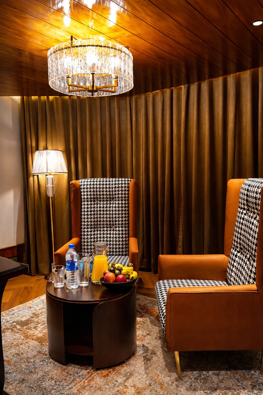 Cozy seating area with two patterned armchairs, a round wooden table holding glasses, a water bottle, orange juice, and a bowl of fruit, set against brown curtains and a wooden ceiling with a chandelier.