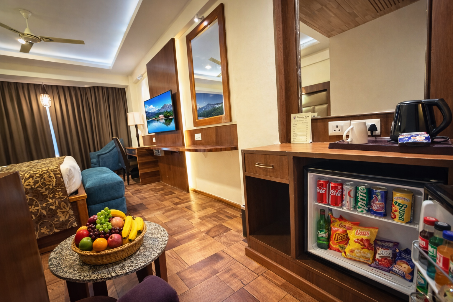 Hotel room with a bed, blue armchair, flat-screen TV, table with a fruit basket, and an open mini-fridge stocked with drinks and snacks.