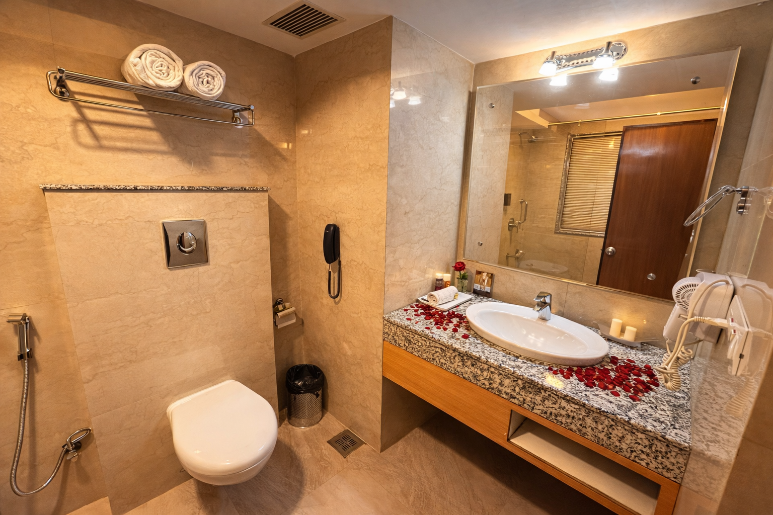 Hotel bathroom with beige marble walls, a wall-mounted toilet, granite countertop with white sink, decorated with rose petals and candles.