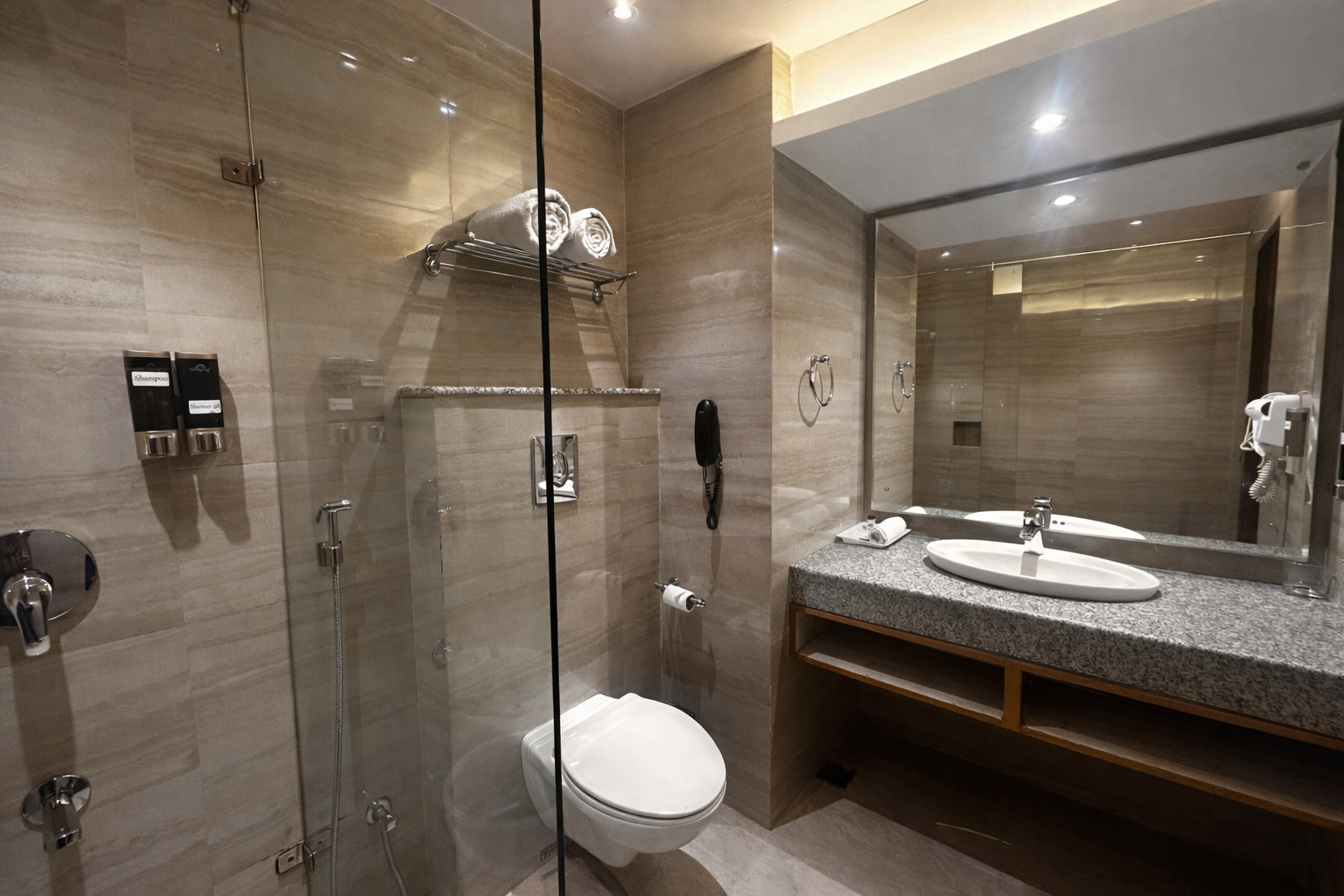 Modern bathroom with a wall-mounted toilet, granite countertop with sink, large mirror, glass shower enclosure, wall-mounted hairdryer, and neatly rolled towels on a rack.