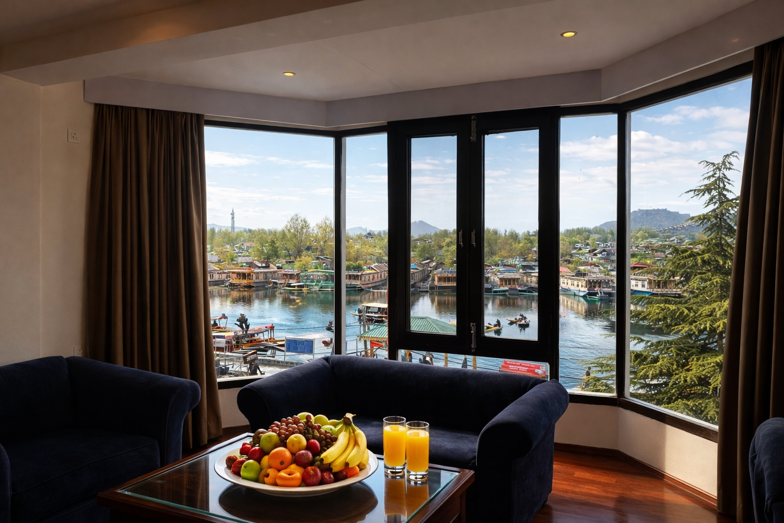 Interior view of a lounge with navy blue sofas, a glass table with a plate of assorted fruits and two glasses of orange juice, and large windows showing a scenic waterfront with boats and greenery.