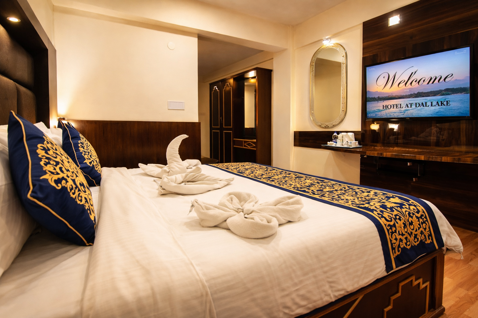 Hotel room with a large bed featuring white linens, blue and gold decorative runner and pillows, towel art, mirror, wardrobe, and a TV displaying a welcome message.