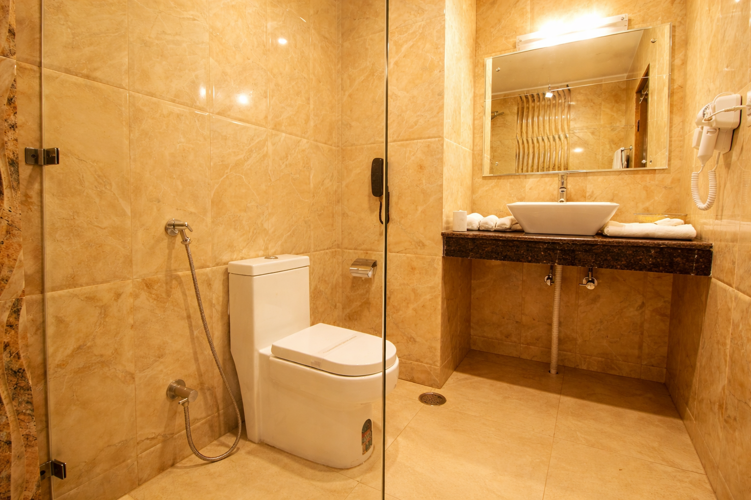 Modern bathroom with beige marble tiles, a white toilet, a sink with countertop, a large mirror, and a wall-mounted hairdryer.