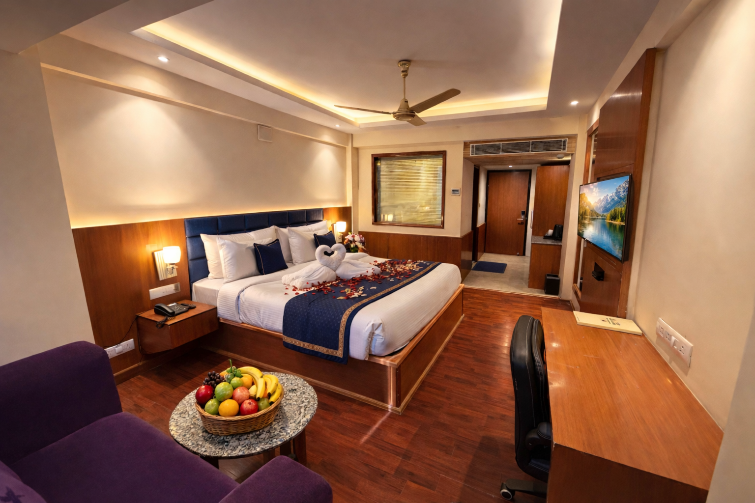 Deluxe hotel room with a large bed decorated with towel swans and rose petals, a fruit basket on a side table, a purple sofa, wooden floor, desk with chair, and a wall-mounted TV.