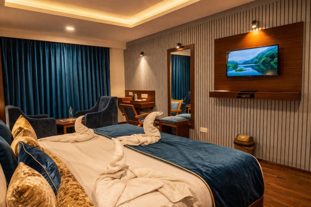 Modern hotel room with a large bed adorned with blue and gold pillows and towel swans, a wall-mounted TV, and cozy seating area with blue curtains.