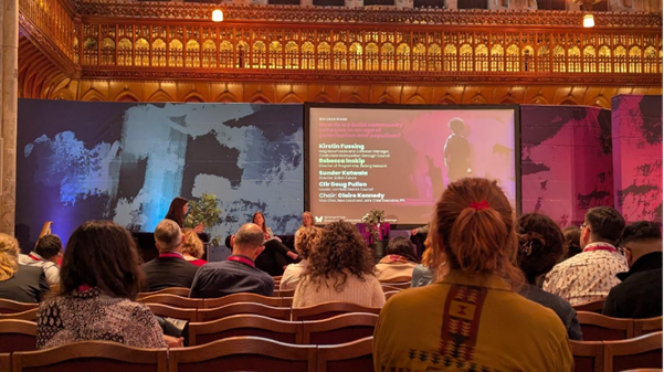 udience seated in the Guild Hall room with an elaborate walls with a stage in front that has a with a large screen displaying speakers' names and details.