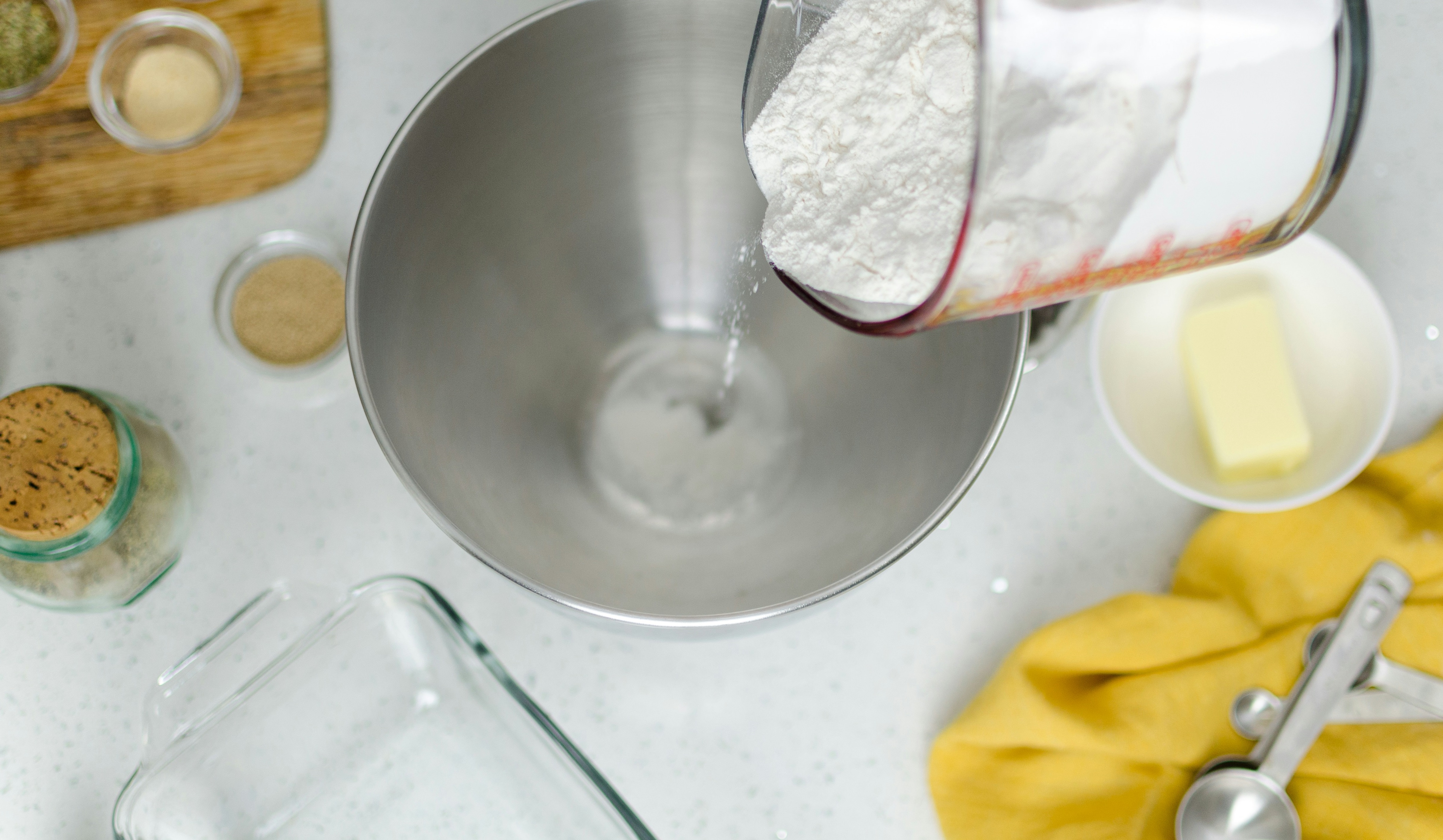 Flour being poured from a measuring cup into a metal mixing bowl on a kitchen counter, surrounded by ingredients including butter, yeast, spices, measuring spoons, and a glass baking dish Photo credit to – Photo credit: Sammee Anderson from Unsplash