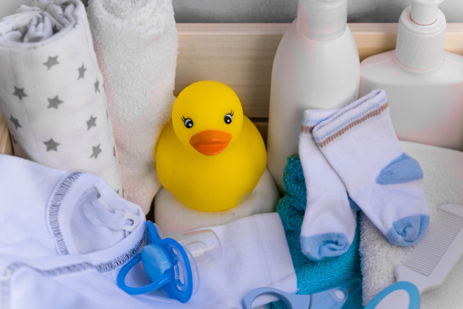 A collection of baby-care items arranged in a wooden box, including a yellow rubber duck in the center, baby lotion bottles, folded towels, a pair of small socks, a pacifier, baby scissors, a comb, and a rolled blanket with star patterns. - Photo Credit Canva Pro