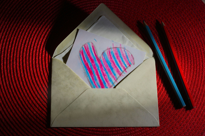 Photo of a white envelop on with a peice of paper in with childs drawing of a heart in pink an dred stripes on a read background with the pink and blue crayons next to it. by Immo Wegmann from Unsplash