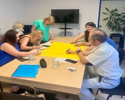 Group photo of 6 members from the Parent2Parent(4 men and 2 women) support programme collaborating together round a pine table writing notes on a big peice of yellow paper, in a white room with a screen and plant in the background.