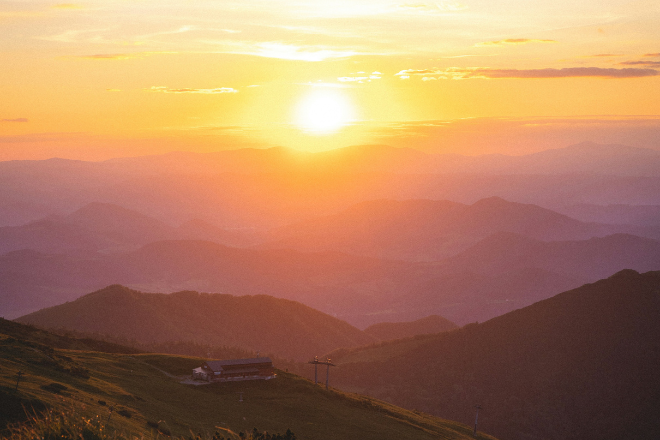 Golden sunset over layered mountain ranges, with rolling hills fading into the distance and a small cabin on a grassy slope in the foreground.