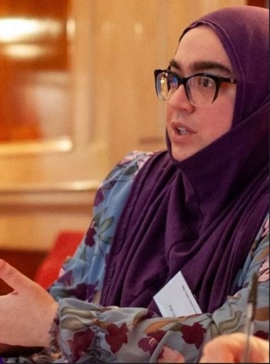 Photo of Aurora who is a woman with glasses wearing a burgundy hijab and a grey and burgundy blouse with flowers on it.