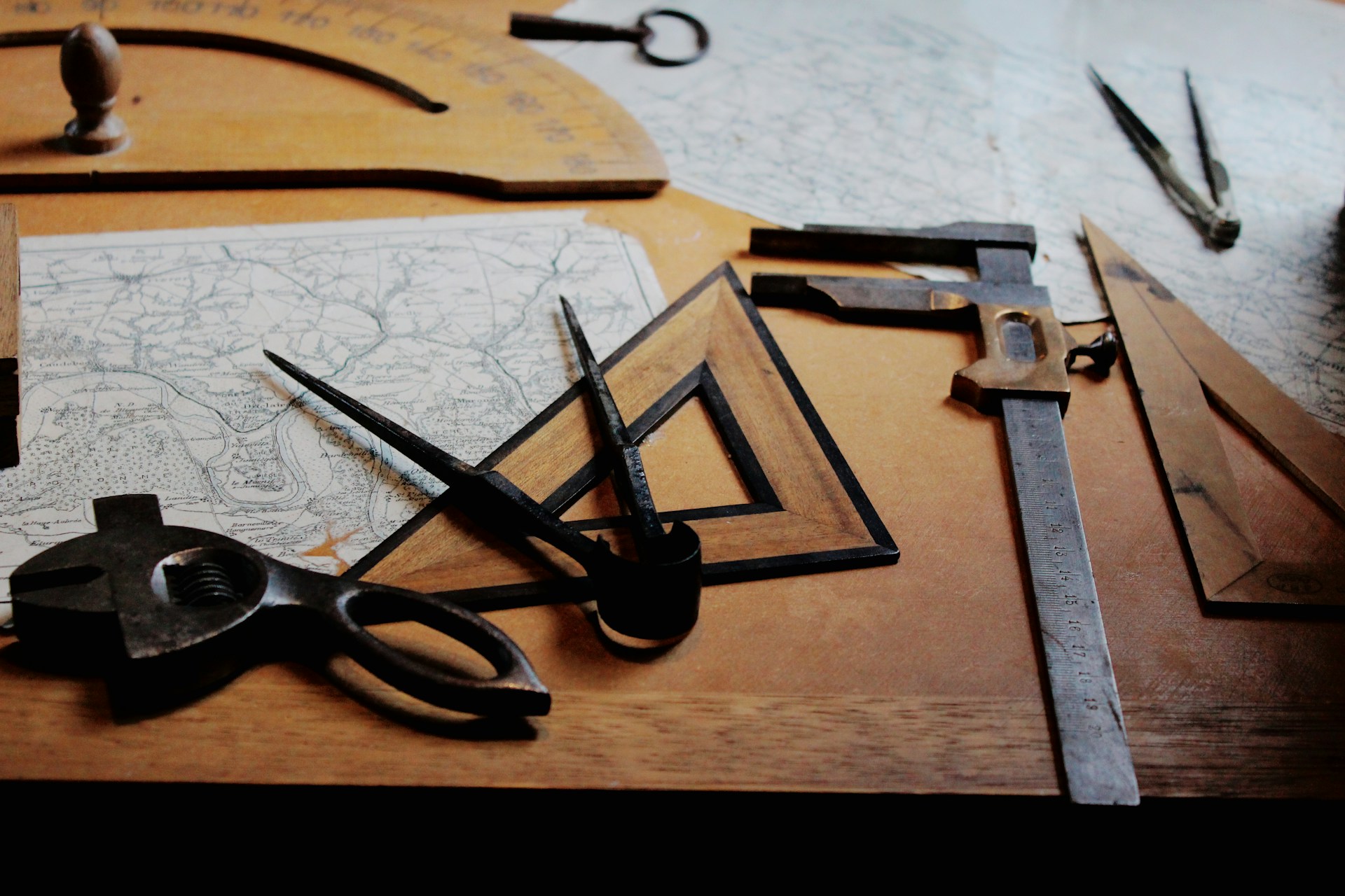 Photo of 2 map on woodent table with a wooden set square and protractor and several compasses scattered around. Photo Credit -Fleur from Unsplash