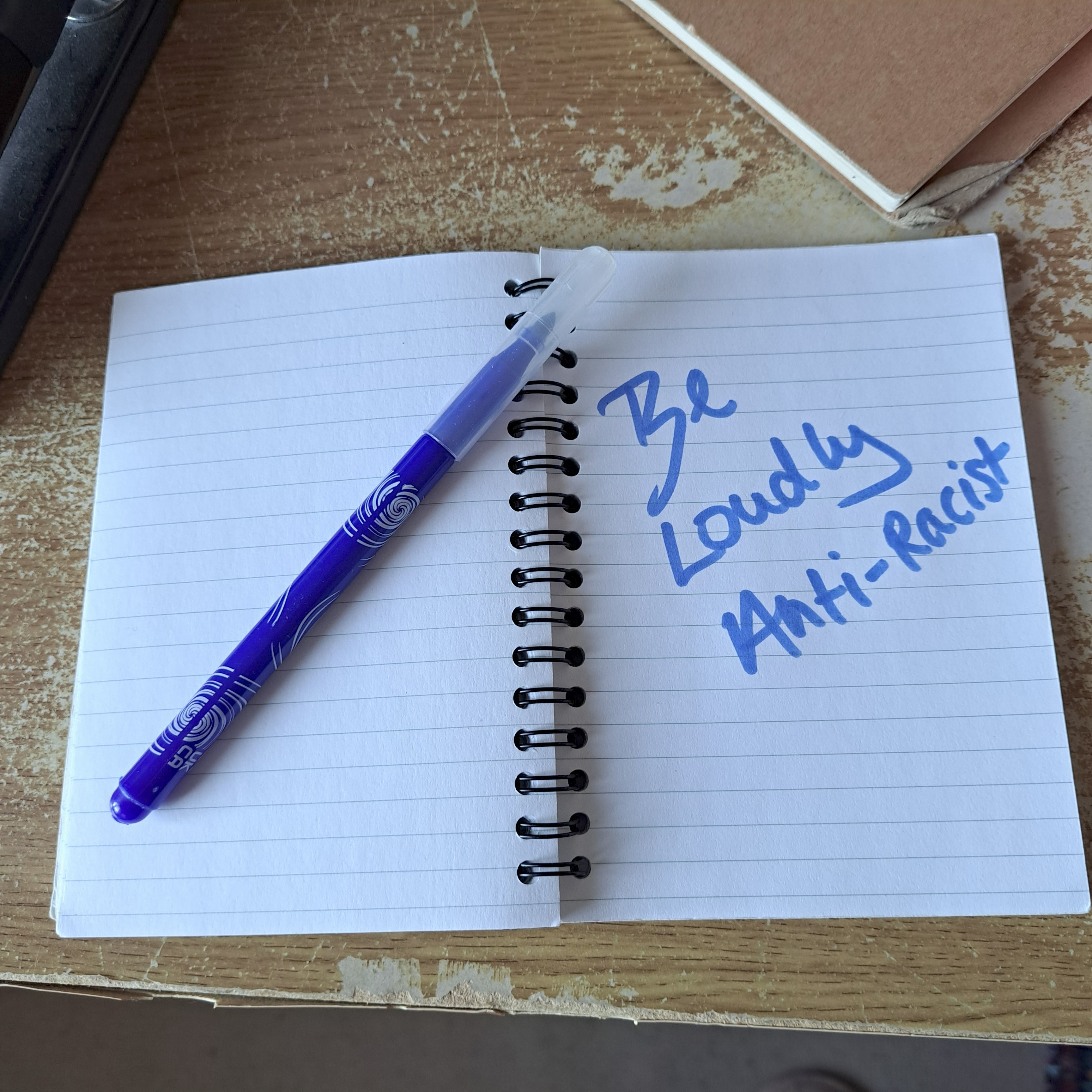 Photo of a wire bound note pad open on a wood desk with a pen resting on one page on and on the opposite page it says 'Be loudly anti-racist' - Photo Credit George Halfin