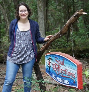 Photo of George a woman wearing jeans and blue top standing in some woods next to sign that says 'Soul Reflections'