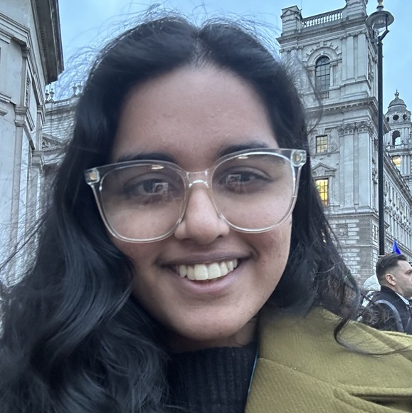 Sindhu is a South Asian woman with dark wavy hair and clear-framed glasses smiling at the camera in an outdoor selfie. They are wearing a dark sweater and an olive-green coat, with historic stone buildings visible in the background at dusk.