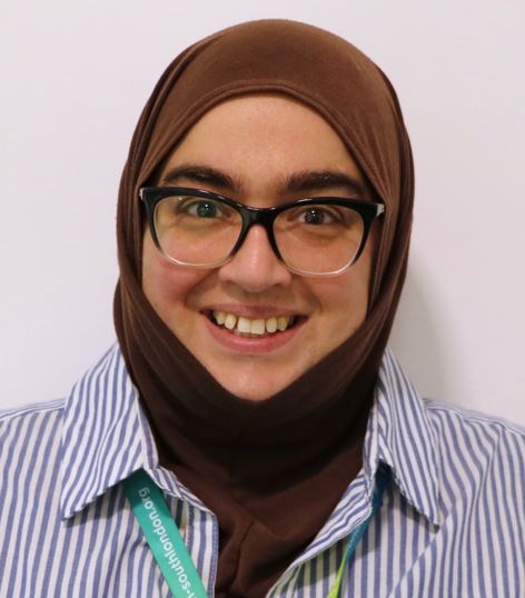 Head-and-shoulders professional portrait of a smiling woman wearing glasses, a brown headscarf, and a blue-and-white striped shirt, looking directly at the camera against a plain light background.
