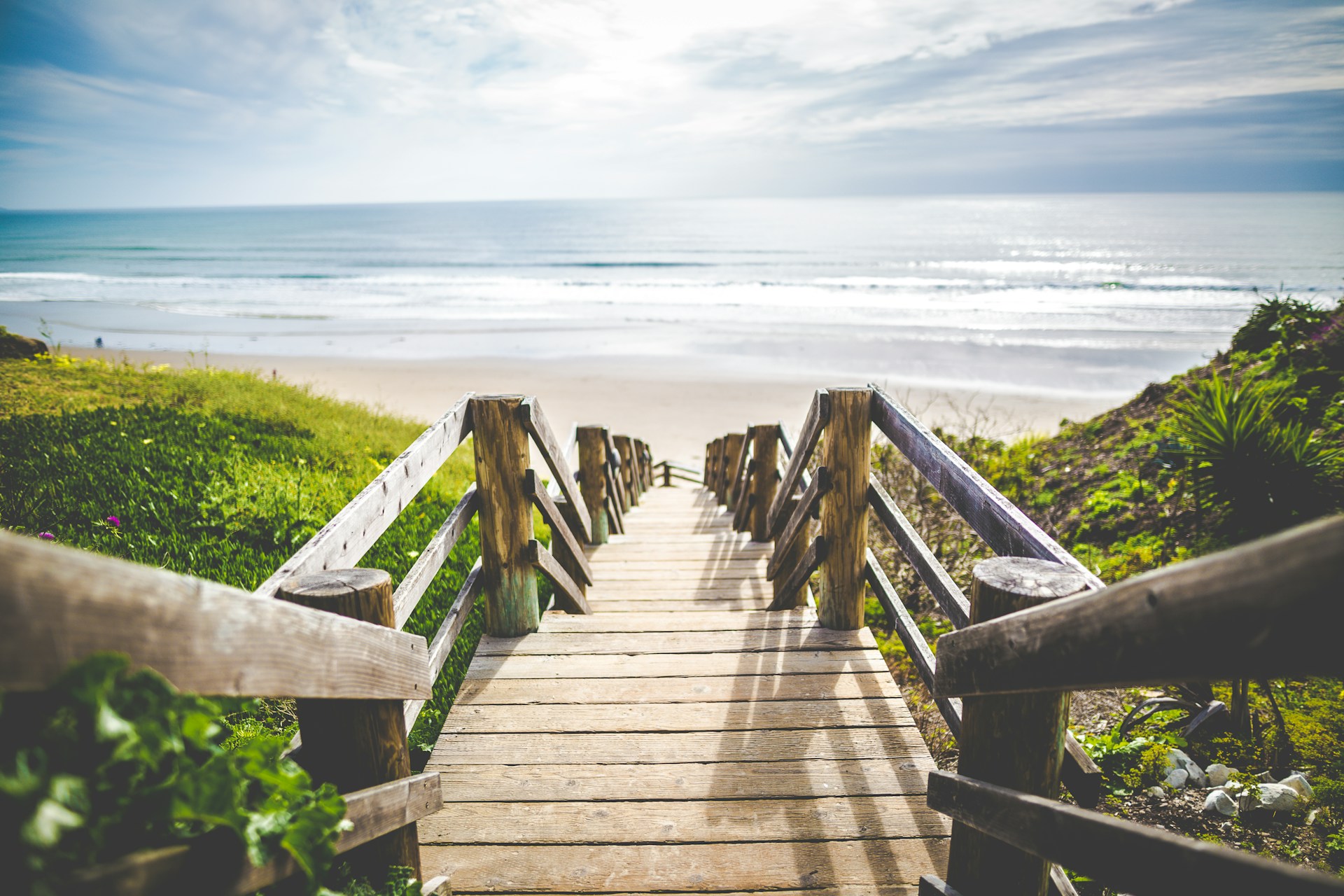 Wooden steps lead down to a beach, with sand dunes and long grass on either side.