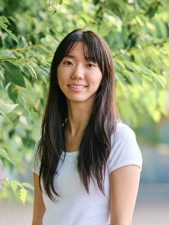 Chaeyeon is an East Asian woman with long dark hair and a fringe, wearing a white T-shirt. She is smiling at the camera, standing in front of green foliage.