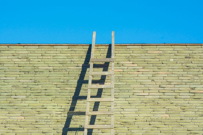 A wooden ladder leaning against a pale brick wall. Blue sky is seen above the wall.