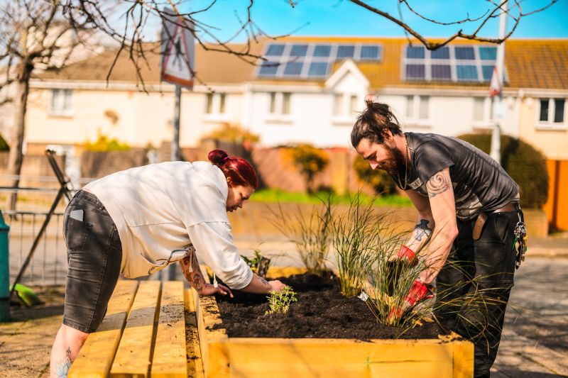 Two people lean over a large new wooden planter planting grasses and bedding plants into the soil, the man on the right is wearing work gloves. In the background is a blurred view of the street.
