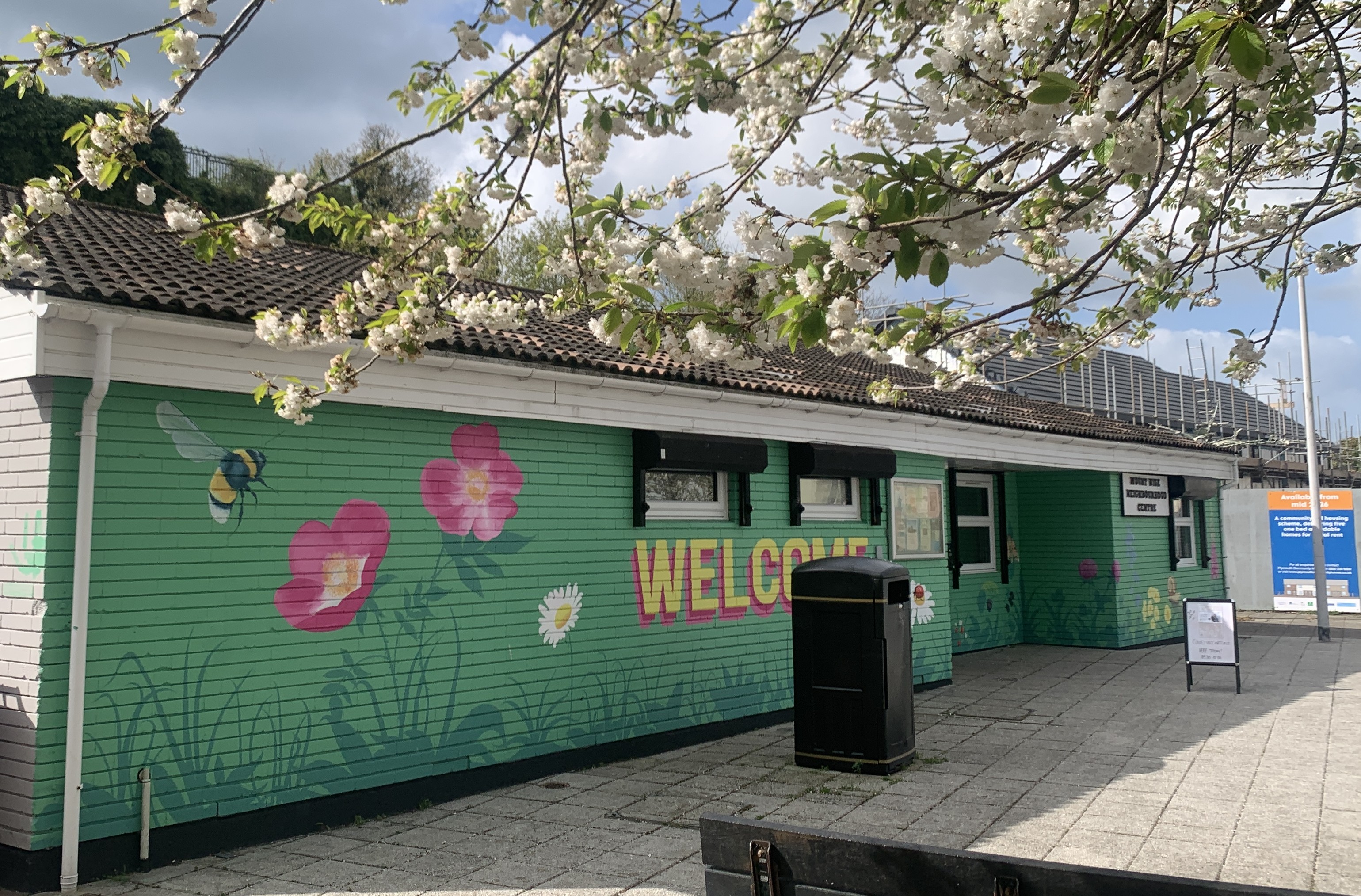 The entire front of a 1960s single-storey building is painted with a mural in light green with a large bumble bee flying over two large pink rose-like flowers, daisies and other greenery. The word WELCOME in large yellow letters with a pink drop shadow is partly obscured by a black bin on the wide pavement in front of the building. Beautiful white blossoms from a tree are seen in the foreground above the building.