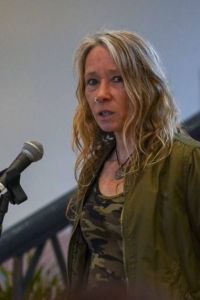 A photo of Debbie, who is speaking into a microphone. Debbie has blonde, shoulder-length hair and is wearing a green jacket.