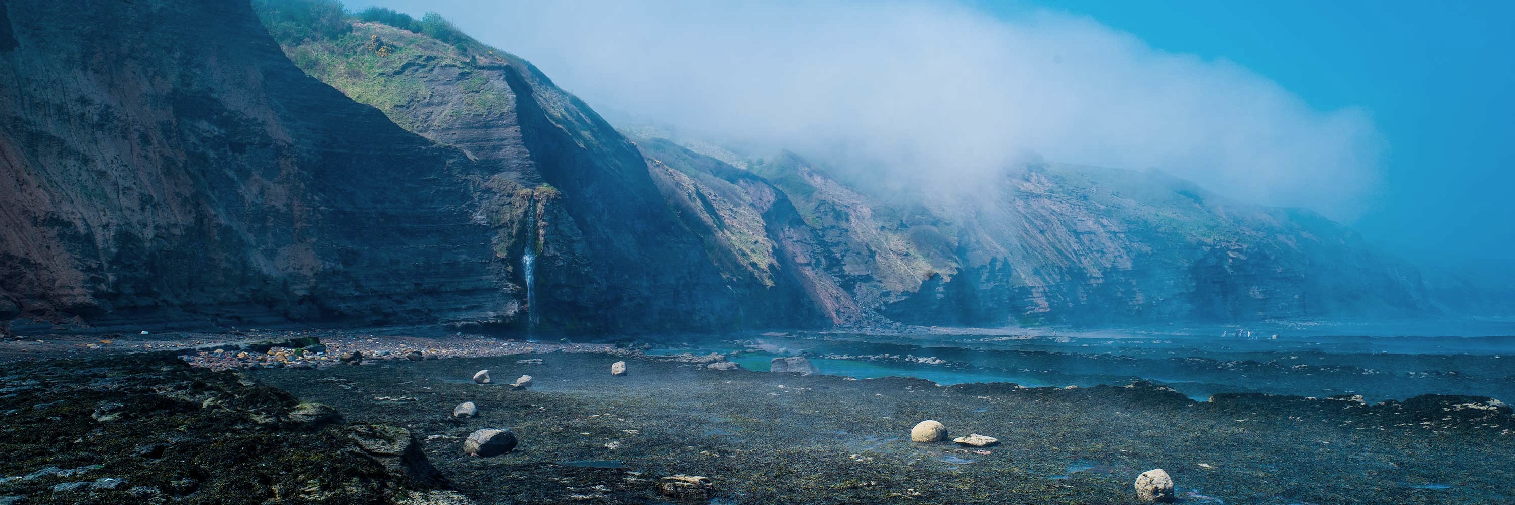 Misty sea cliffs with a waterfall above a rocky shore at low tide
