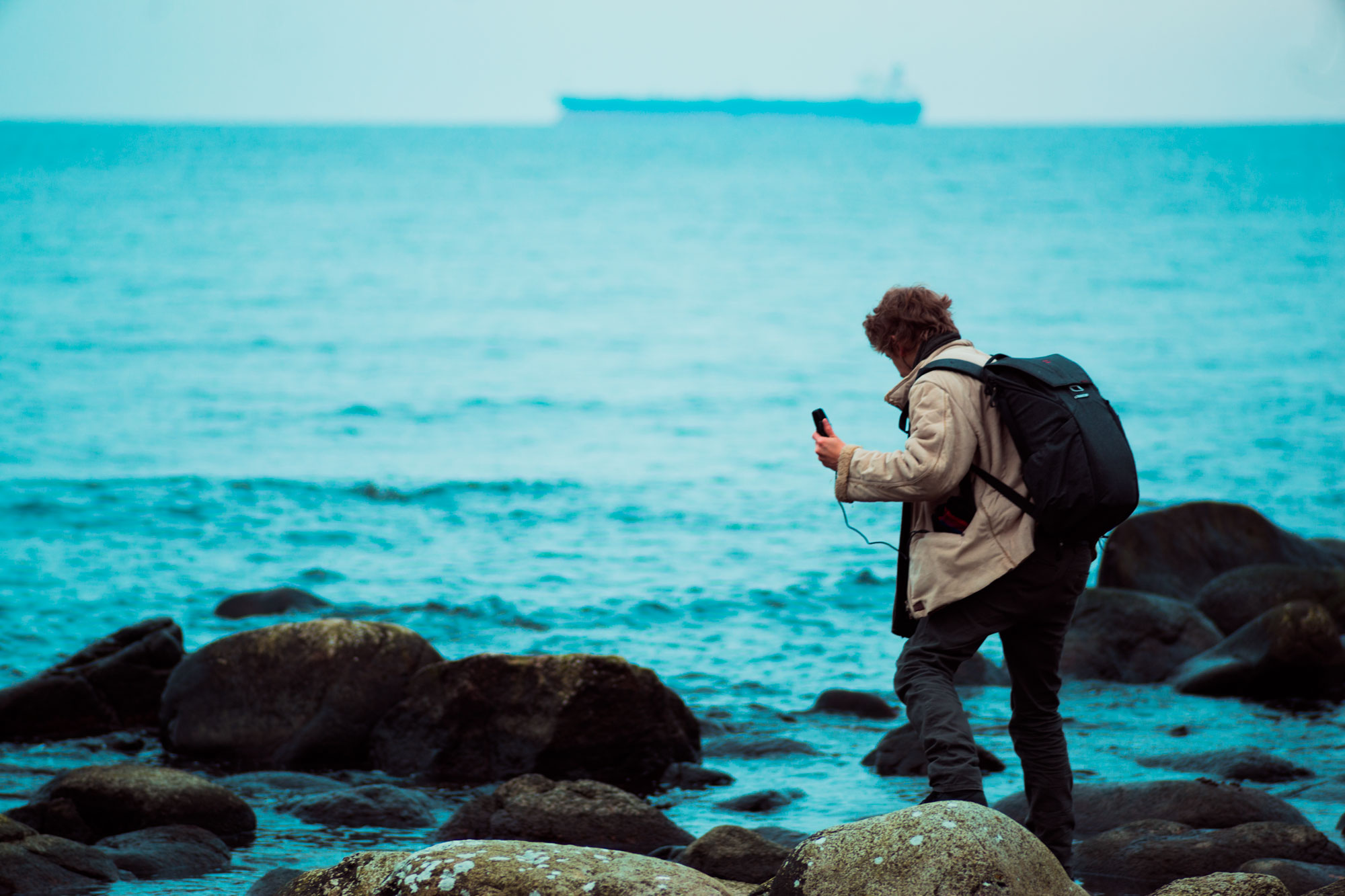 Markus recording sound with a handheld recorder on a rocky shore, a ship on the far horizon
