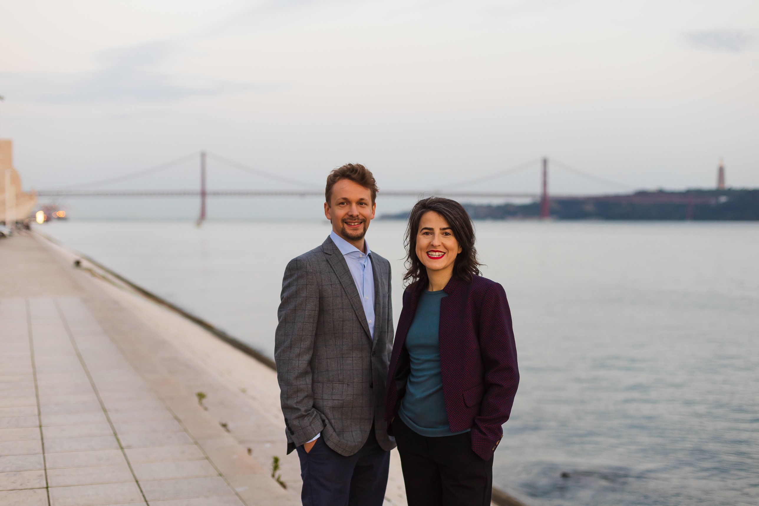 Markus Pesonen and Catarina Brazão on the Lisbon waterfront at dusk, the 25 de Abril bridge behind them