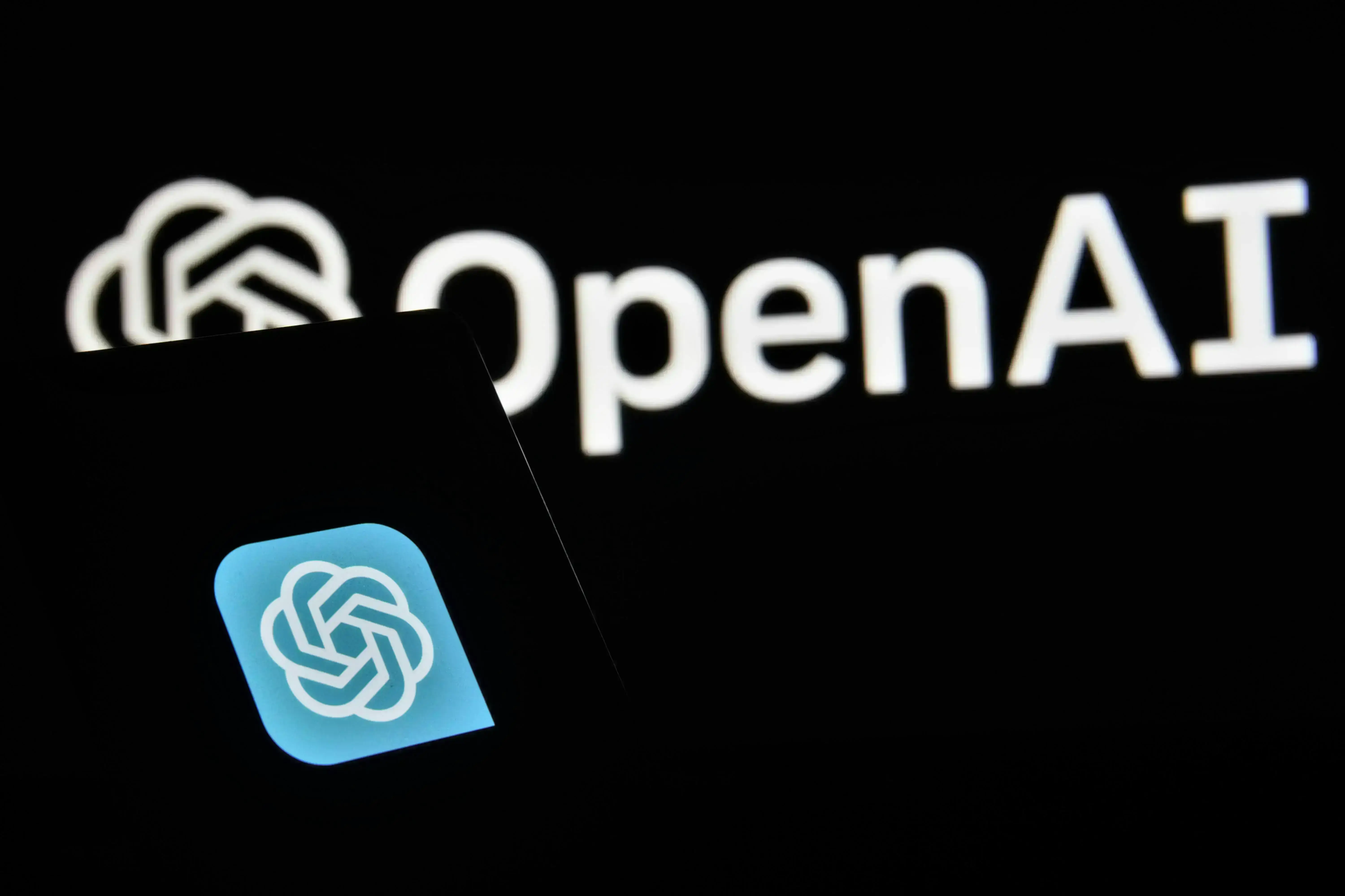 A smartphone screen displays the OpenAI logo in blue, overlapping a blurred, larger version of the AI-driven OpenAI logo behind it