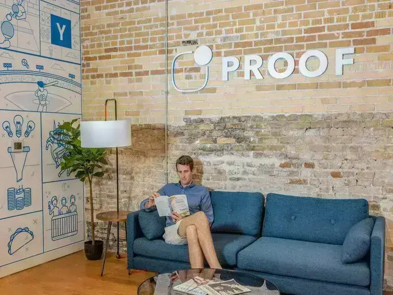 A man is sitting on a blue couch in a modern office with exposed brick walls, reading a book about digital marketing.