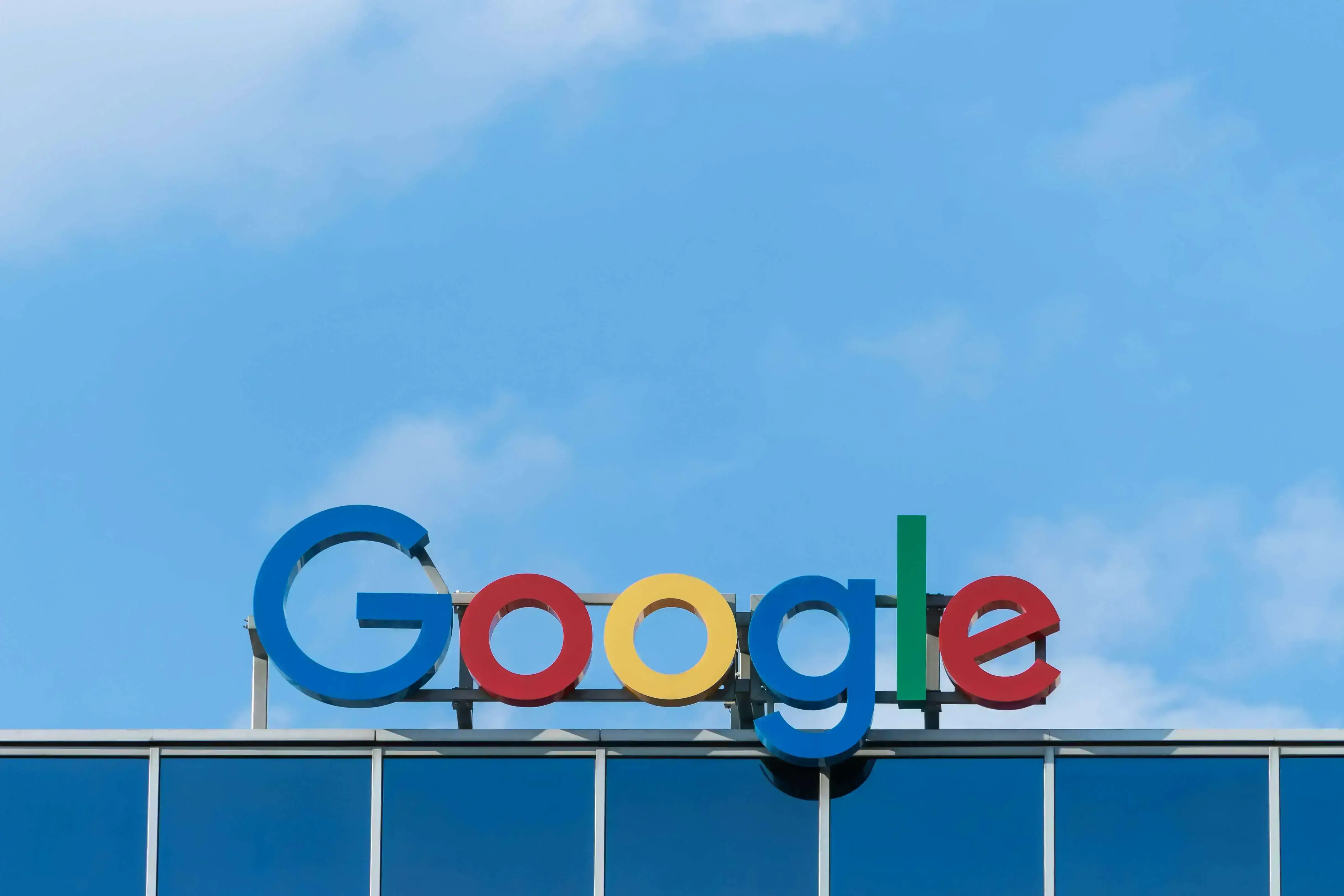 A large, colorful Google logo is displayed on the exterior of a modern glass building against a backdrop of a clear blue sky with a few clouds.