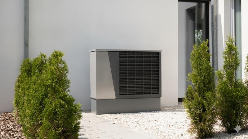 An air source heat pump unit mounted on the exterior wall of a modern white home, surrounded by neatly landscaped shrubs.