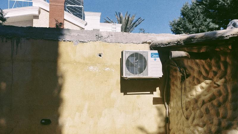 A heat pump unit mounted on a sun-baked exterior wall with solar panels visible on the rooftop above against a clear blue sky.