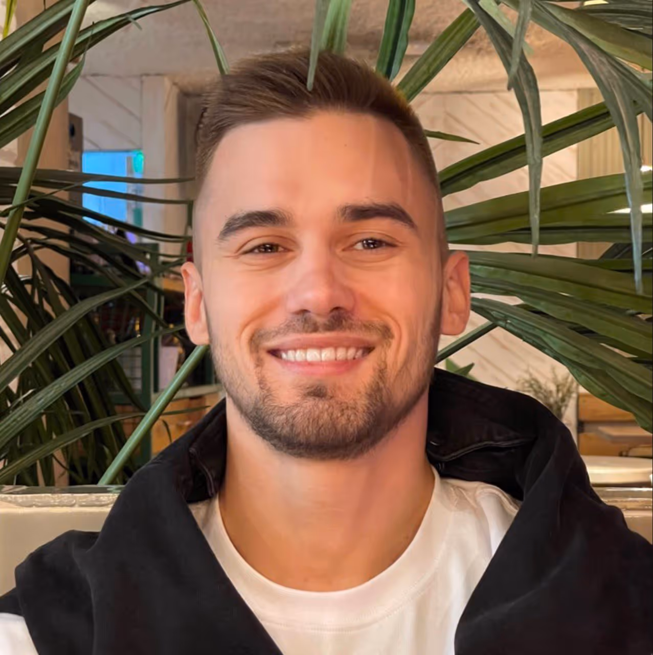 Smiling young man with short hair and a trimmed beard wearing a white shirt and black jacket, sitting indoors with green plants in the background.