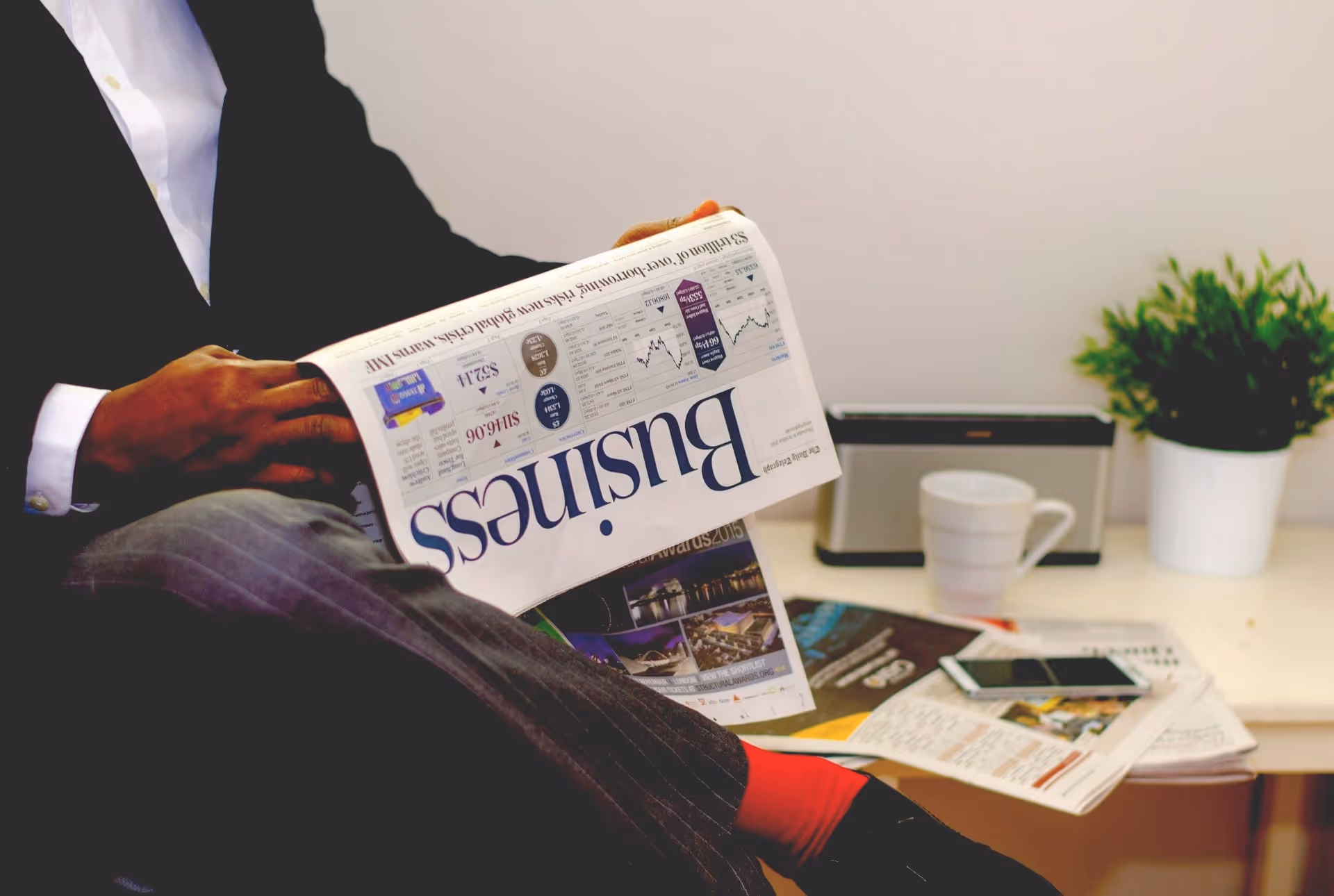 Person in business attire reading a business newspaper with red socks visible, sitting next to a table with a cup, smartphone, and plant.